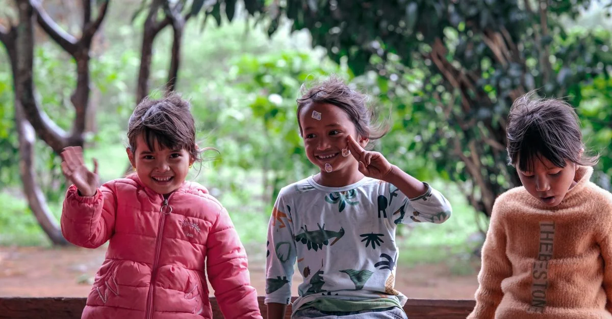 Trois enfants joyeux sont assis sur un banc dans un cadre extérieur luxuriant. Ils sourient et interagissent joyeusement.